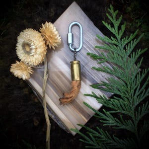 May include: A unique keychain featuring a brass bullet casing and a small, brown, furry object. The keychain is attached to a silver carabiner clip. The background includes dried flowers, a wooden surface, and green foliage.