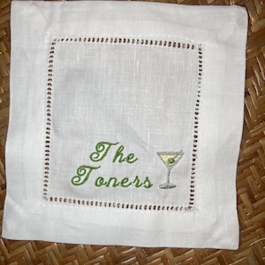 May include: White linen cocktail napkin with a decorative stitched border. The napkin features green embroidered text that reads "The Toners" and a martini glass with an olive.