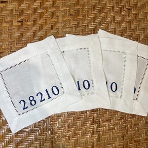 May include: A set of four white linen cocktail napkins with a stitched border. Each napkin has a navy blue embroidered number. The first napkin displays the number "28210". The napkins are arranged on a woven, light brown surface.
