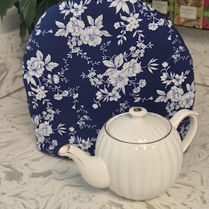May include: A white ceramic teapot with vertical ribbing and gold trim sits in front of a dark blue fabric tea cozy featuring a white floral pattern. The teapot has a matching lid with a gold knob, and the tea cozy has a small fabric loop at the top.