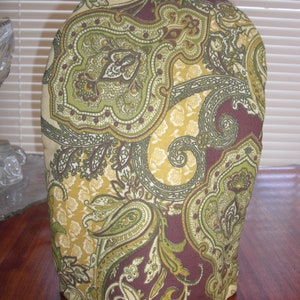May include: A fabric tea cozy featuring a detailed paisley pattern in shades of olive green, mustard yellow, and deep burgundy. The dome-shaped cover has a small fabric loop at the top center and a rounded base, designed to fit over a teapot.