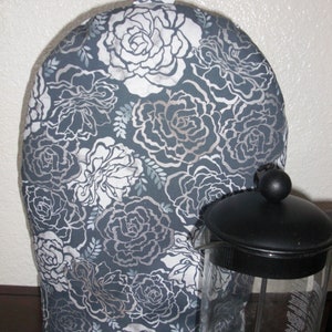 May include: A fabric tea cozy featuring a dark gray background with a repeating white and light gray floral pattern of roses and leaves. The tea cozy has a rounded top with a small fabric loop handle and sits beside a glass French press.
