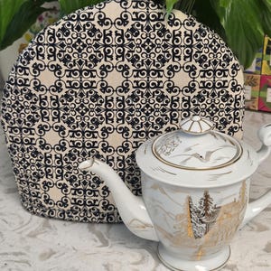 May include: A white porcelain teapot with gold-toned landscape illustrations and a matching lid, positioned in front of a rounded fabric tea cozy featuring a repeating black and cream-colored geometric pattern with a small black fabric tab at the top.