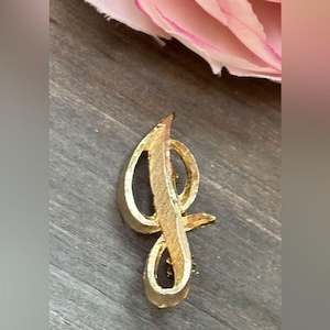 May include: A gold-coloured metal brooch shaped like the letter "J". The brooch features a cursive design with a looped top and a curved base. Set against a dark wood surface with a pink flower.