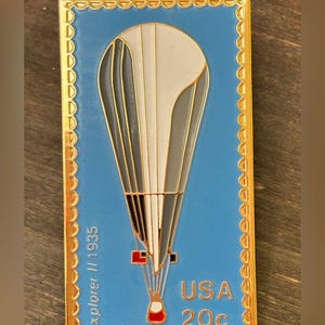 May include: A rectangular enamel pin featuring a light blue background with a gold scalloped border. The design depicts a white and grey hot air balloon with the text Explorer II 1935 on the left and USA 20c on the bottom right corner.