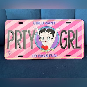May include: Pink license plate with a diagonal stripe pattern and the words "PRTY GRL". It features a Betty Boop graphic and the text "GIRLS WANT TO HAVE FUN!"