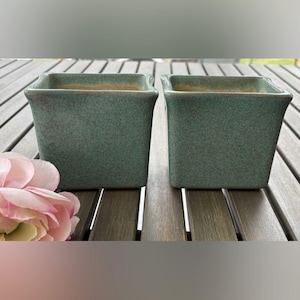May include: Two square, teal-colored ceramic planters with a textured finish. The planters are placed side-by-side on a wooden surface, with a pink flower in the foreground. The planters have a slightly flared rim.