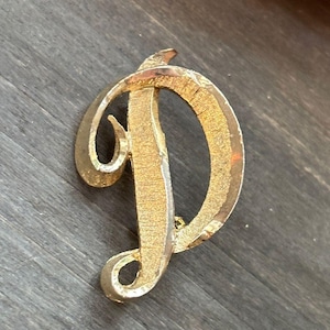May include: Gold-toned metal brooch in the shape of the letter "D". The brooch has a textured surface and a pin closure on the back. The brooch is set against a dark wood grain background.