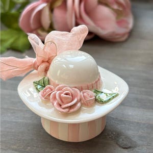 May include: A ceramic hat-shaped trinket box. The hat is white with a pink band and adorned with pink roses and green leaves. A pink ribbon is tied on top. The base is striped pink and white.