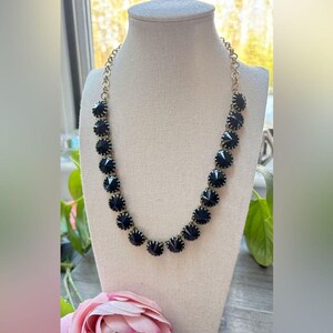 May include: A necklace featuring a series of round, faceted black stones set in gold-toned metal bezels. The stones are linked together in a single strand, creating a circular design that hangs from a gold-toned chain with circular links.