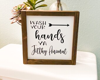 May include: A wooden framed sign with the text "Wash Your Hands Ya Filthy Animal" in black script. The sign is on a white toilet, with a vase of cotton stems and greenery in the background.