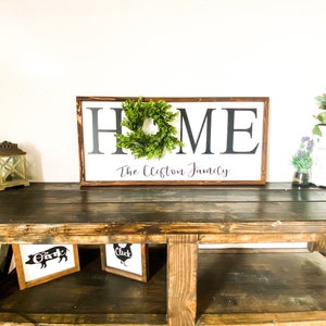 May include: A wooden sign with the word "HOME" in large, black letters, a green wreath, and the family name below. The sign is on a dark wooden table with framed farm animal art and a decorative lantern.