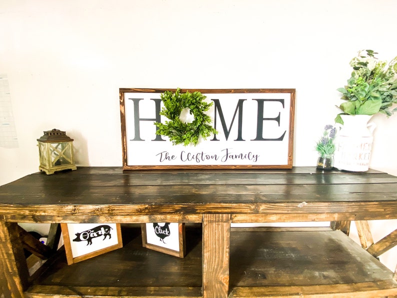 May include: A wooden sign with the word "HOME" in large, black letters, a green wreath, and the family name below. The sign is on a dark wooden table with framed farm animal art and a decorative lantern.