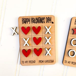 May include: A wooden Valentine's Day card with a tic-tac-toe game. The game board has red hearts and white X's. The card says "Happy Valentines Day." and "To my friend from: Kayleigh".