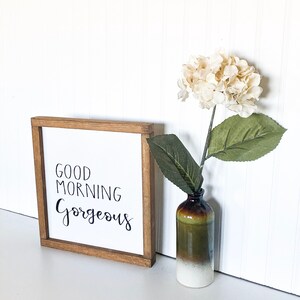 May include: A wooden framed sign with the text "Good Morning Gorgeous" in black script. The sign is sitting on a white surface next to a small vase with a white hydrangea.