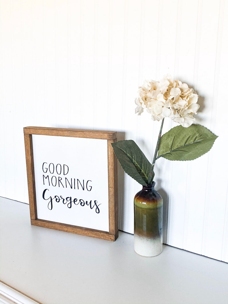 May include: A wooden framed sign with the text "Good Morning Gorgeous" in black script. The sign is sitting on a white surface next to a small vase with a white hydrangea.