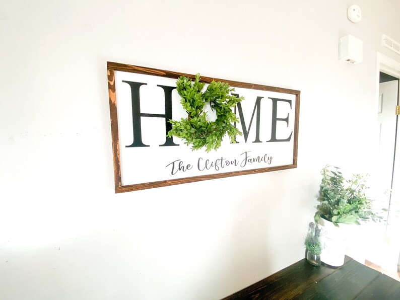 May include: A rectangular wooden sign with the word "HOME" in large black letters, a small green wreath, and the family name "The Clifton Family" in script. The sign is mounted on a white wall.
