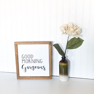 May include: A wooden framed sign with the text "Good Morning Gorgeous" in black script. The sign is sitting on a white surface with a white flower in a brown and white vase.