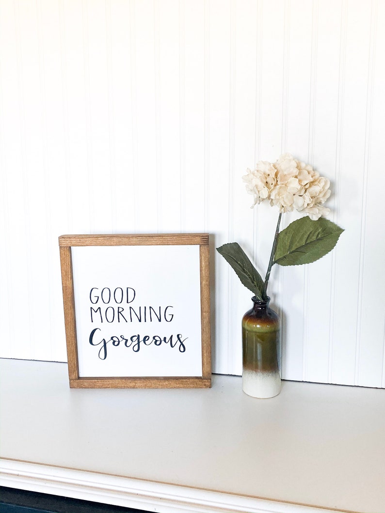 May include: A wooden framed sign with the text "Good Morning Gorgeous" in black script. The sign is sitting on a white surface with a white flower in a brown and white vase.