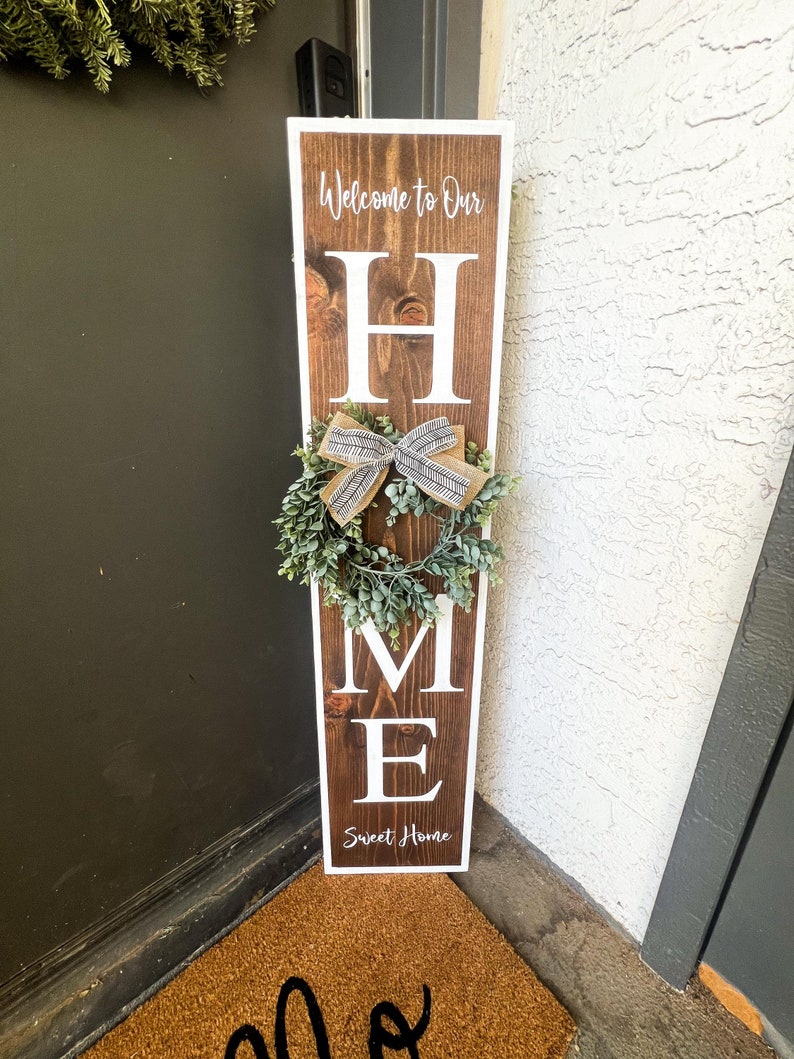 May include: A wooden sign with the word "HOME" in white letters. The sign is decorated with a green wreath and a brown and white bow. The sign also says "Welcome to Our" and "Sweet Home".
