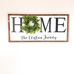 May include: A rectangular wooden sign with the word "HOME" in large black letters on a white background. A small green wreath is attached to the "O". The text "The Clifton Family" is written in cursive below.