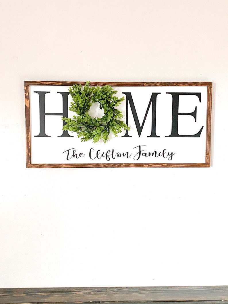 May include: A rectangular wooden sign with the word "HOME" in large black letters on a white background. A small green wreath is attached to the "O". The text "The Clifton Family" is written in cursive below.
