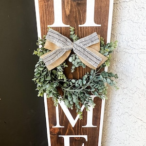 May include: A wooden sign with the word "HOME" in white letters. The sign is decorated with a small wreath of green leaves and a brown and white bow. The text "Sweet Home" is written at the bottom of the sign.
