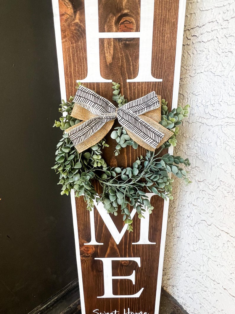 May include: A wooden sign with the word "HOME" in white letters. The sign is decorated with a small wreath of green leaves and a brown and white bow. The text "Sweet Home" is written at the bottom of the sign.