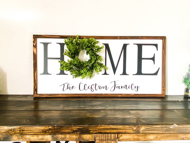 May include: A rectangular wooden sign with a white background and the word "HOME" in large black letters. A small green wreath is centered on the "O". Below the word "HOME" is the text "The Clifton Family" in a script font.
