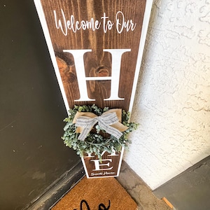 May include: A wooden sign with the words "Welcome to Our Home" and a large white letter "H" in the centre. The sign is decorated with a small green wreath with a brown and white ribbon. The sign is leaning against a door with a brown doormat that says "Hello" in black.