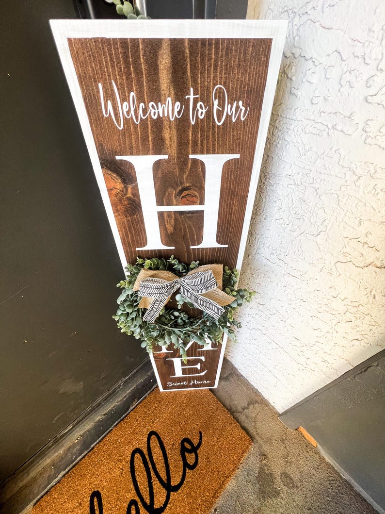May include: A wooden sign with the words "Welcome to Our Home" and a large white letter "H" in the centre. The sign is decorated with a small green wreath with a brown and white ribbon. The sign is leaning against a door with a brown doormat that says "Hello" in black.