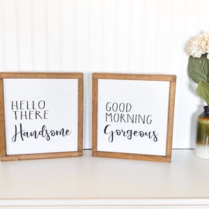 May include: Two framed signs with white backgrounds and black lettering. The sign on the left says "Hello There Handsome" and the sign on the right says "Good Morning Gorgeous".