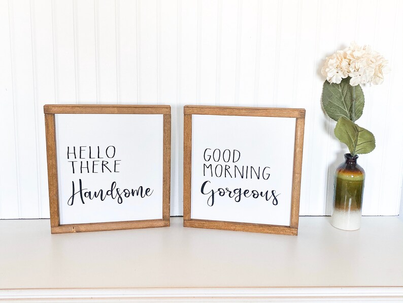 May include: Two framed signs with white backgrounds and black lettering. The sign on the left says "Hello There Handsome" and the sign on the right says "Good Morning Gorgeous".