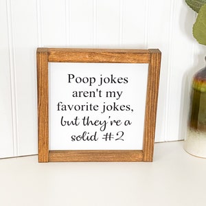May include: A brown wooden framed sign with white background and black text that reads "Poop jokes aren't my favorite jokes, but they're a solid #2"
