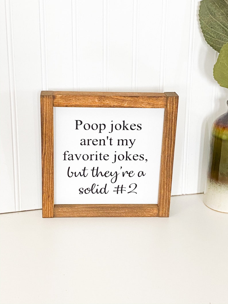 May include: A brown wooden framed sign with white background and black text that reads "Poop jokes aren't my favorite jokes, but they're a solid #2"