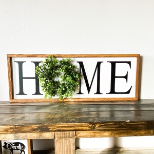 May include: A rectangular wooden sign with the word "HOME" in bold black letters on a white background. A small green wreath is centered above the "O". The sign is displayed on a rustic wooden table, with holiday decorations in the background.
