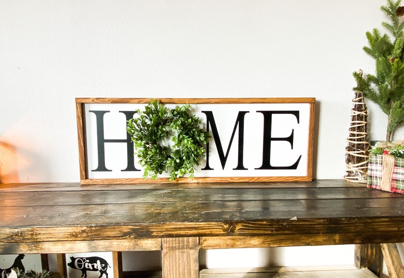 May include: A rectangular wooden sign with the word "HOME" in bold black letters on a white background. A small green wreath is centered above the "O". The sign is displayed on a rustic wooden table, with holiday decorations in the background.