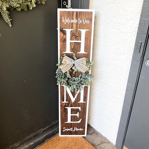May include: A wooden sign with the word "HOME" in white letters. The sign is decorated with a small wreath and a bow. The sign also says "Welcome to Our" and "Sweet Home".