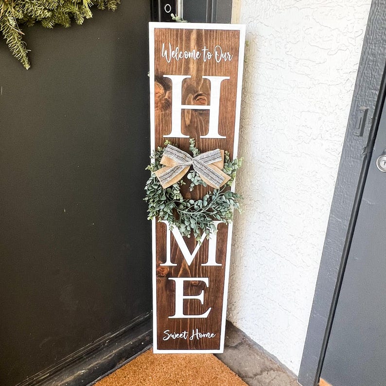 May include: A wooden sign with the word "HOME" in white letters. The sign is decorated with a small wreath and a bow. The sign also says "Welcome to Our" and "Sweet Home".