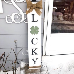 May include: A white wooden sign with the word "LUCKY" spelled out in black letters. The sign is decorated with a burlap bow and a green shamrock.