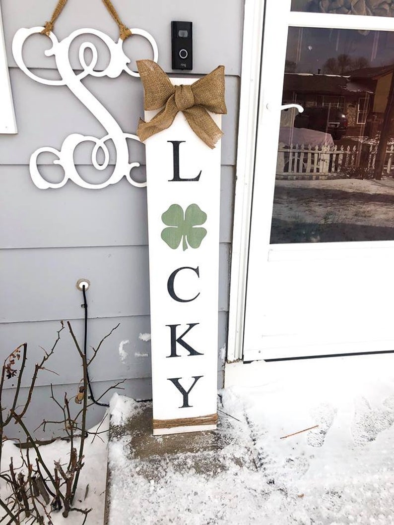 May include: A white wooden sign with the word "LUCKY" spelled out in black letters. The sign is decorated with a burlap bow and a green shamrock.