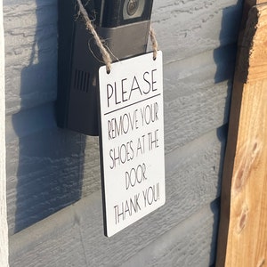 May include: A white rectangular sign with the text "PLEASE REMOVE YOUR SHOES AT THE DOOR. THANK YOU!" is hanging from a black security camera. The sign is attached with twine and is mounted on a gray wall.