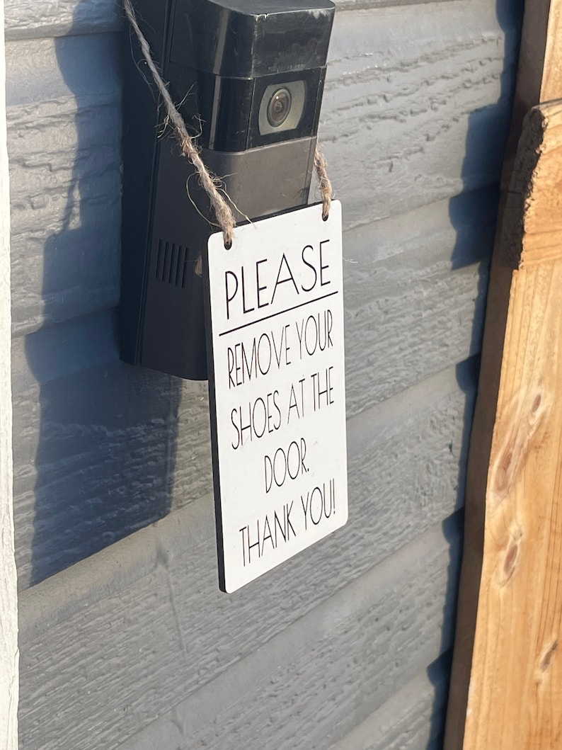 May include: A white rectangular sign with the text "PLEASE REMOVE YOUR SHOES AT THE DOOR. THANK YOU!" is hanging from a black security camera. The sign is attached with twine and is mounted on a gray wall.