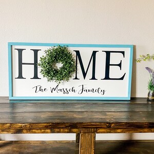 May include: A rectangular wooden sign with a light blue frame. The word "HOME" is in large black letters, with a small green wreath in the center of the "O". Below, in script, is "The Massich Family".