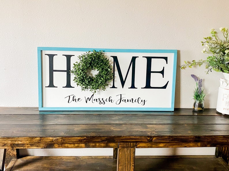 May include: A rectangular wooden sign with a light blue frame. The word "HOME" is in large black letters, with a small green wreath in the center of the "O". Below, in script, is "The Massich Family".