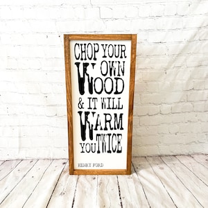 May include: Wooden framed sign with the quote "CHOP YOUR OWN WOOD & IT WILL WARM YOU TWICE" in black text on a white background. The quote is attributed to Henry Ford. The frame is a dark brown color.