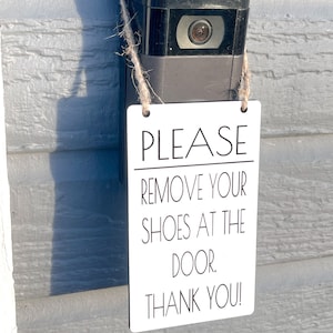 May include: A black video doorbell with a white rectangular sign that reads "PLEASE REMOVE YOUR SHOES AT THE DOOR. THANK YOU!" The sign is suspended by twine.
