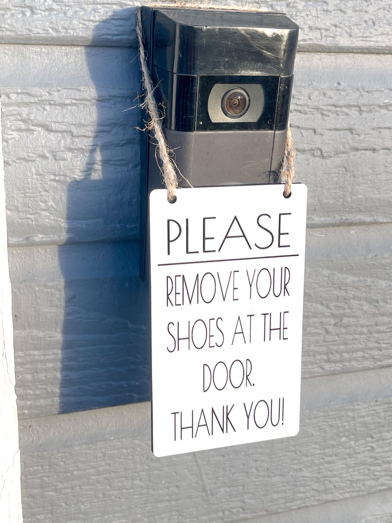 May include: A black video doorbell with a white rectangular sign that reads "PLEASE REMOVE YOUR SHOES AT THE DOOR. THANK YOU!" The sign is suspended by twine.