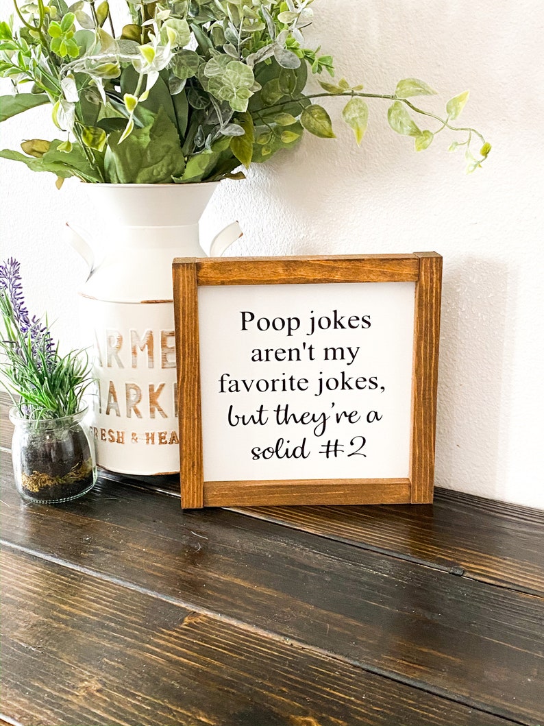 May include: A wooden framed sign with white background and black text that reads "Poop jokes aren't my favorite jokes, but they're a solid #2".