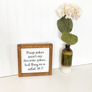 May include: A wooden sign with the text "Poop jokes aren't my favorite jokes, but they're a solid #2". The sign is sitting on a white surface with a white flower in a brown and green vase.